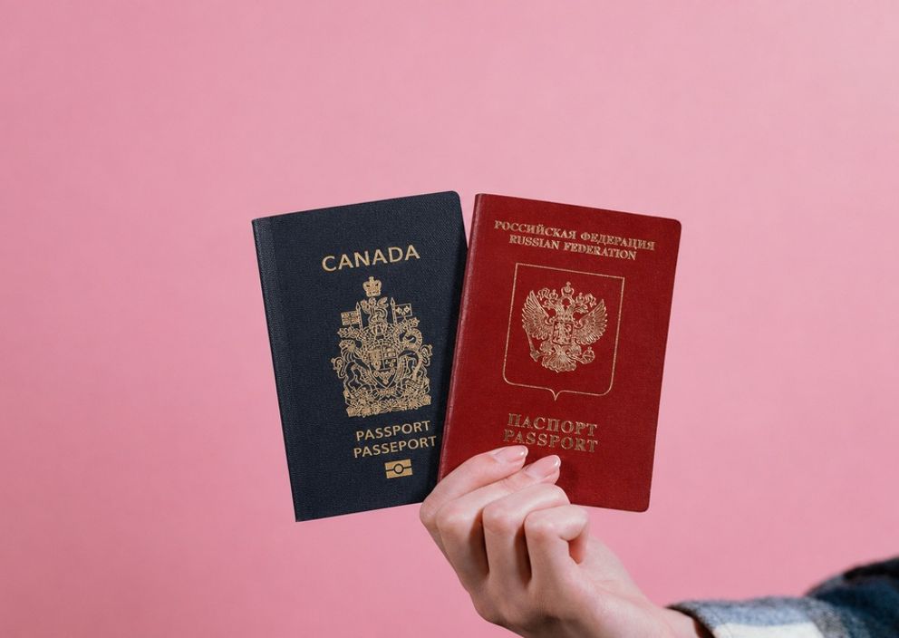 A hand holding a Canadian passport and a Russian passport side-by-side against a pink background.