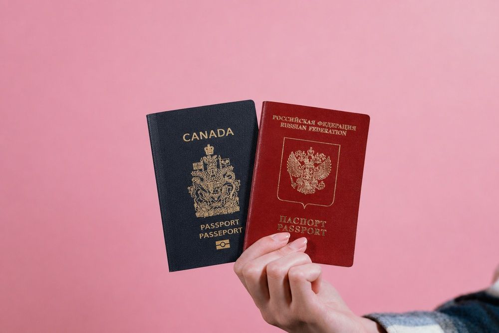 A hand holding a Canadian passport and a Russian passport side-by-side against a pink background.