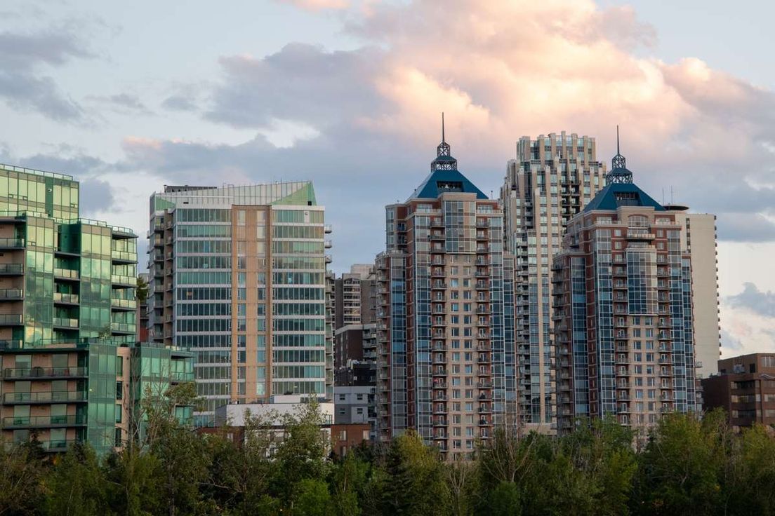 High-rise modern buildings in an Alberta city rising above green trees under a cloudy sky at dusk.