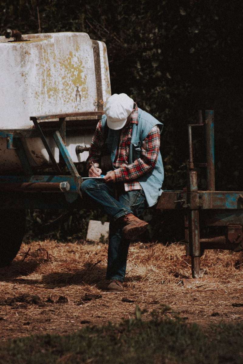 A man in a plaid shirt and cap sits on farm equipment writing on a clipboard.