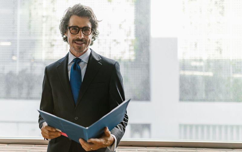 A man wearing a dark suit, tie, and glasses stands near a window holding an open blue folder.