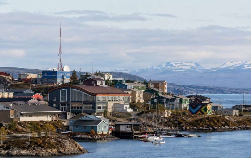 A northern town with colorful buildings, floatplanes at a dock, and snow-capped mountains across the water.