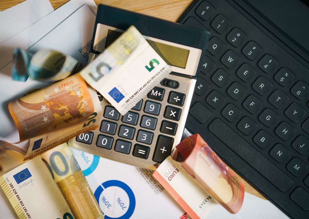 Euro banknotes, a calculator, financial charts, and a laptop keyboard on a desk.