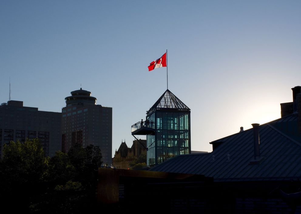 A Canadian flag flying atop a modern glass tower against a clear sky in a city skyline.