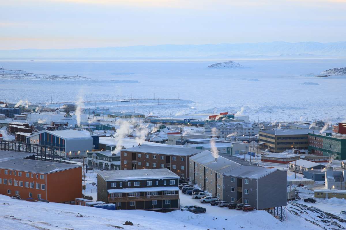A snow-covered town in Nunavut with smoke rising from buildings beside a frozen bay.
