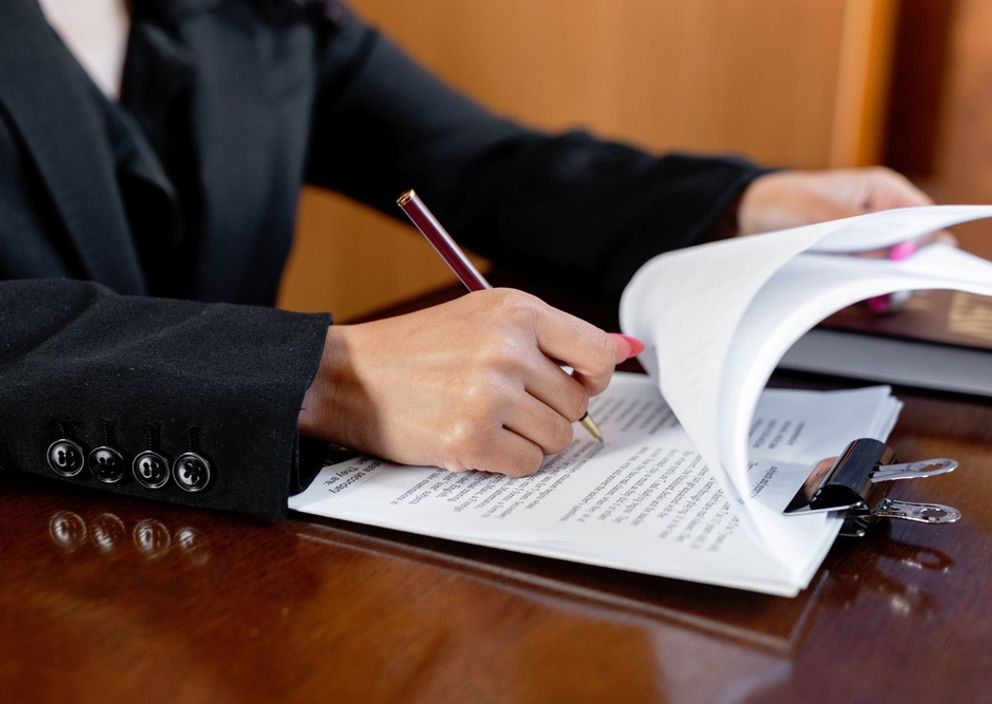 Close-up of a person in a black suit reviewing and signing documents on a clipboard at a desk.