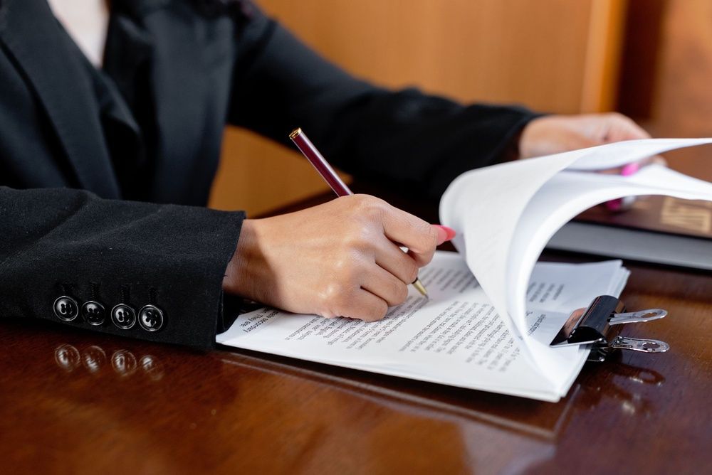 Close-up of a person in a black suit reviewing and signing documents on a clipboard at a desk.