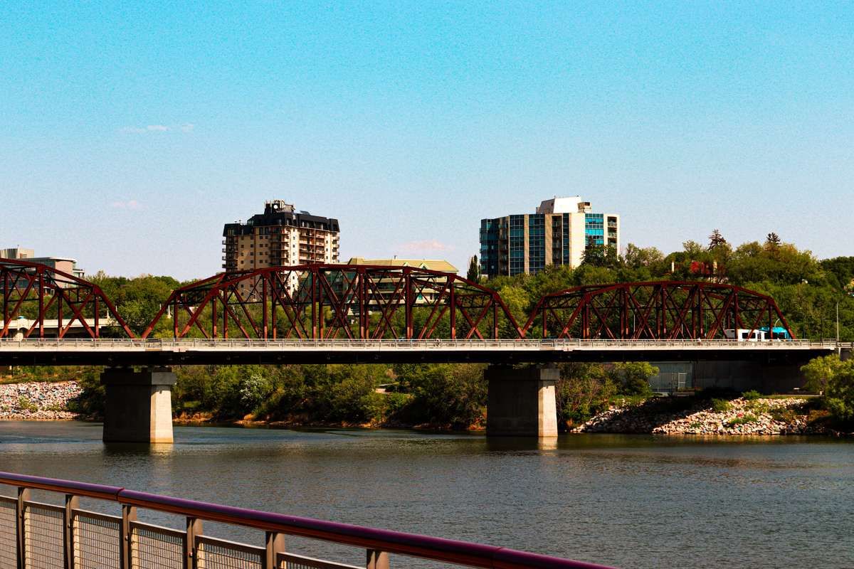 A red steel truss bridge crossing a river with city buildings and trees in the background under a blue sky.