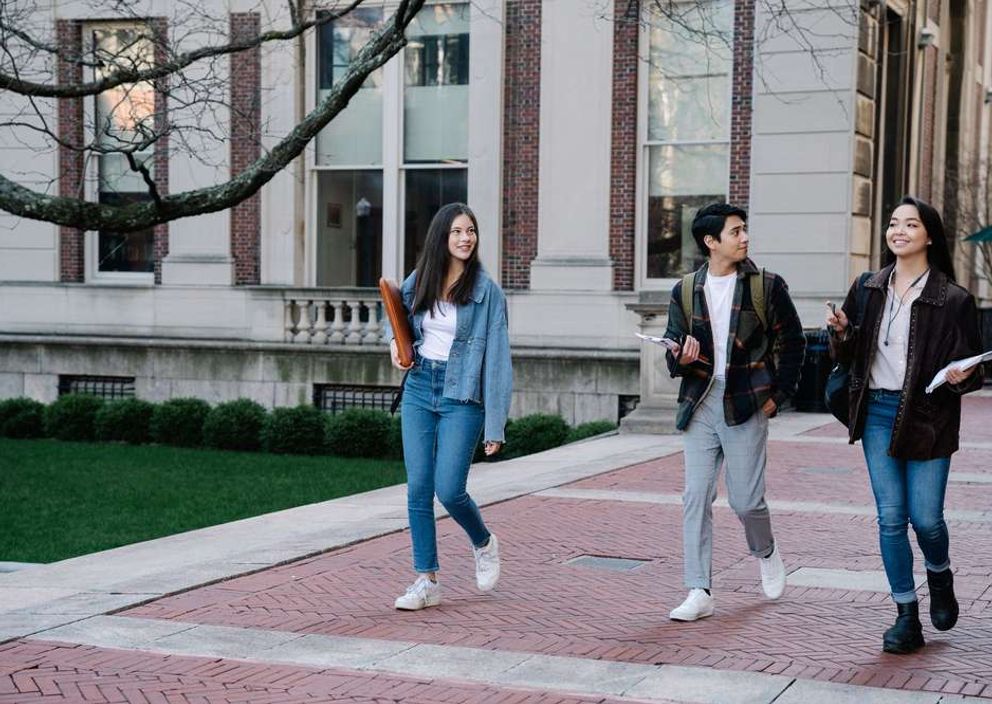 Three university students walk and chat on a brick pathway outside an academic building.