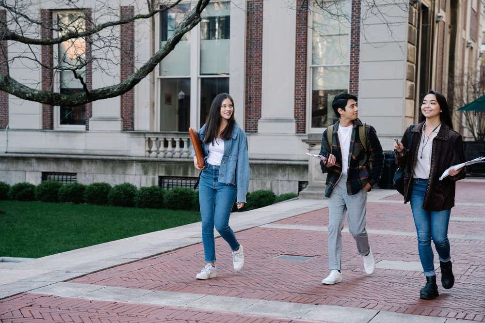 Three university students walk and chat on a brick pathway outside an academic building.